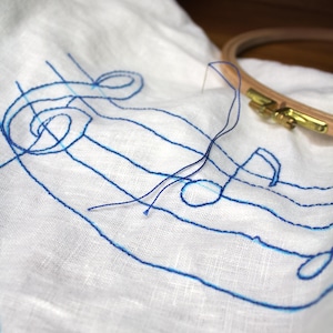 May include: A white linen fabric featuring a blue embroidered treble clef and musical staff lines. A thin blue thread extends from the embroidery, and a wooden embroidery hoop with a brass tension screw rests on the fabric near the design.