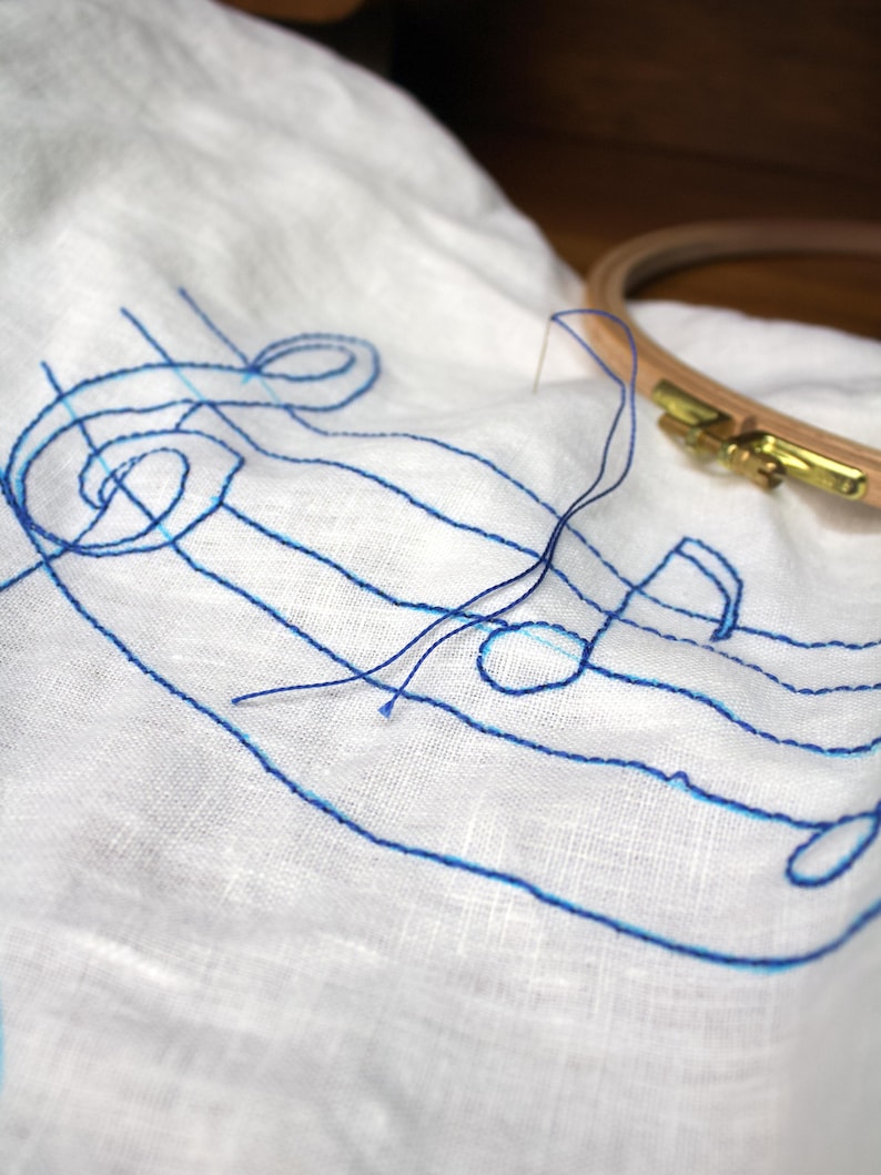 May include: A white linen fabric featuring a blue embroidered treble clef and musical staff lines. A thin blue thread extends from the embroidery, and a wooden embroidery hoop with a brass tension screw rests on the fabric near the design.