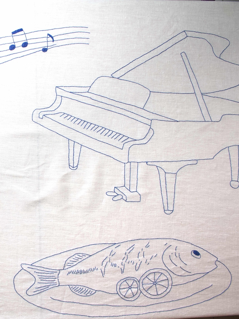 Detailed shot of the embroidery: blue musical staff with notes and a grand piano outline on a white linen background.