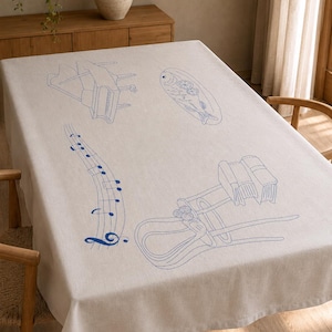 May include: A rectangular beige tablecloth featuring blue embroidered line art illustrations. The designs include a grand piano, a fish on a platter, a musical staff with notes, and a wooden chair with a stack of books resting on the seat.