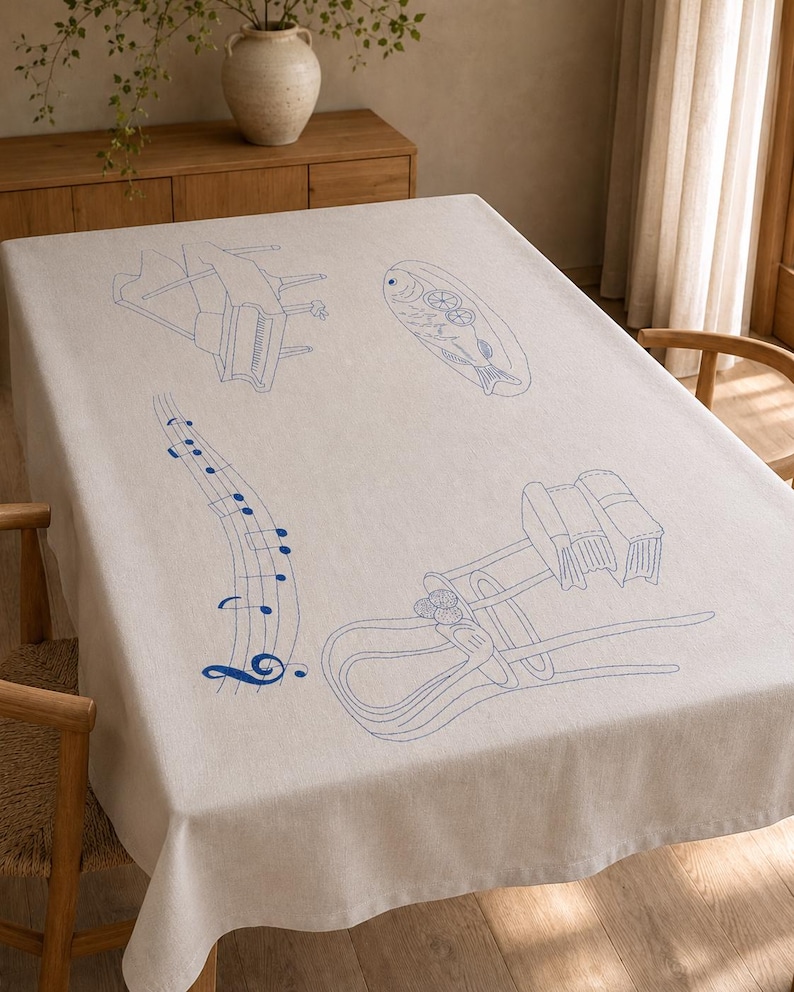 May include: A rectangular beige tablecloth featuring blue embroidered line art illustrations. The designs include a grand piano, a fish on a platter, a musical staff with notes, and a wooden chair with a stack of books resting on the seat.