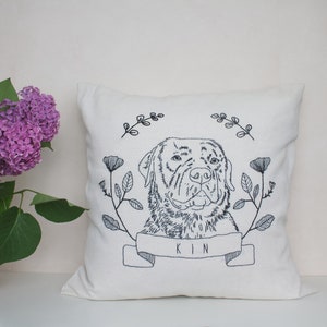 May include: Cream-colored square throw pillow with an embroidered dog portrait. The dog is surrounded by floral and leaf designs, with the word "KIN" embroidered on a banner below the dog's face. The embroidery is in dark blue.