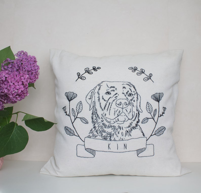 May include: Cream-colored square throw pillow with an embroidered dog portrait. The dog is surrounded by floral and leaf designs, with the word "KIN" embroidered on a banner below the dog's face. The embroidery is in dark blue.