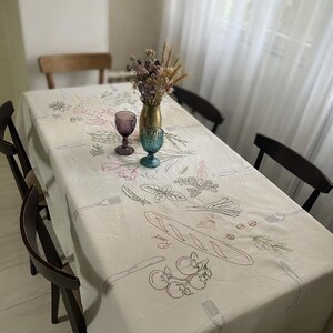 May include: A rectangular off-white tablecloth featuring embroidered line art illustrations of food items including a baguette, tomatoes, herbs, and cutlery. The fabric is draped over a dining table surrounded by four dark wooden chairs.