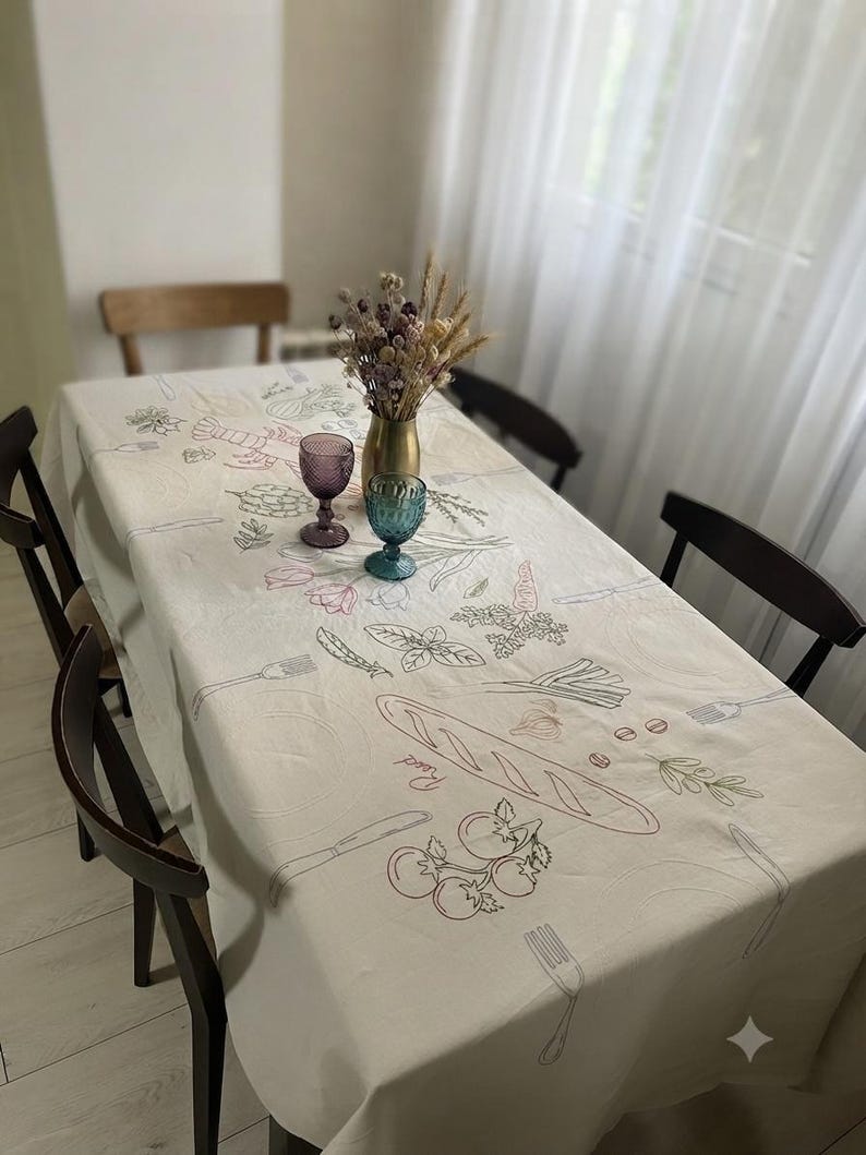 May include: A rectangular off-white tablecloth featuring embroidered line art illustrations of food items including a baguette, tomatoes, herbs, and cutlery. The fabric is draped over a dining table surrounded by four dark wooden chairs.