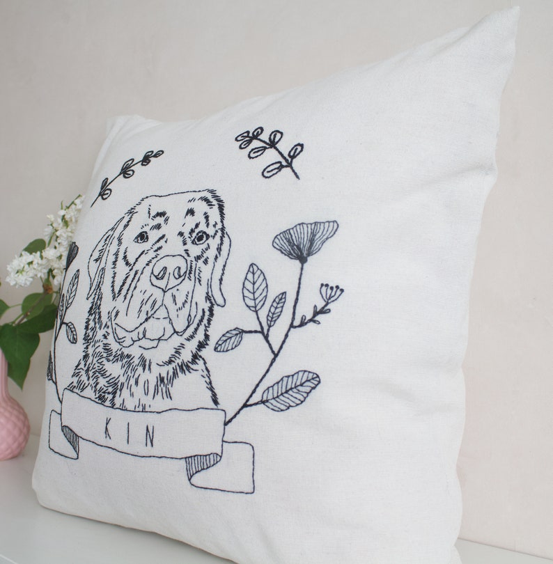 May include: Cream-colored square pillow with a detailed black embroidered design. The design features a dog's portrait, floral elements, and the word "KIN" on a banner. The pillow is made of a textured fabric.