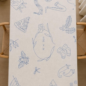 May include: A rectangular off-white tablecloth featuring blue line art illustrations including a saxophone, pizza slice, bee, baby socks, chili peppers, martini glass, and a central oval frame containing the date 15 11 2025 and text SE24.