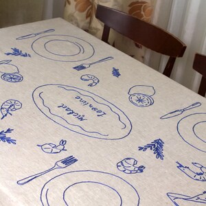 May include: A beige linen tablecloth featuring blue embroidered designs of plates, forks, knives, shrimp, lemons, and sprigs. A central oval motif contains the embroidered names Michael and Leontine in a cursive script, creating a dining theme.
