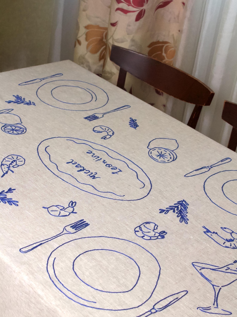 May include: A beige linen tablecloth featuring blue embroidered designs of plates, forks, knives, shrimp, lemons, and sprigs. A central oval motif contains the embroidered names Michael and Leontine in a cursive script, creating a dining theme.