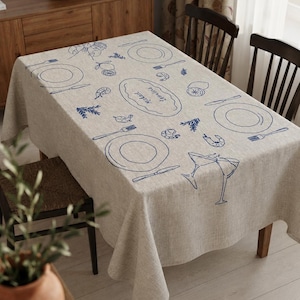 May include: A rectangular beige linen tablecloth featuring a blue line-art print of place settings, shrimp, lemons, herbs, and two clinking cocktail glasses. A central oval shape contains the handwritten names "Mickael" and "Leontine" in blue ink.