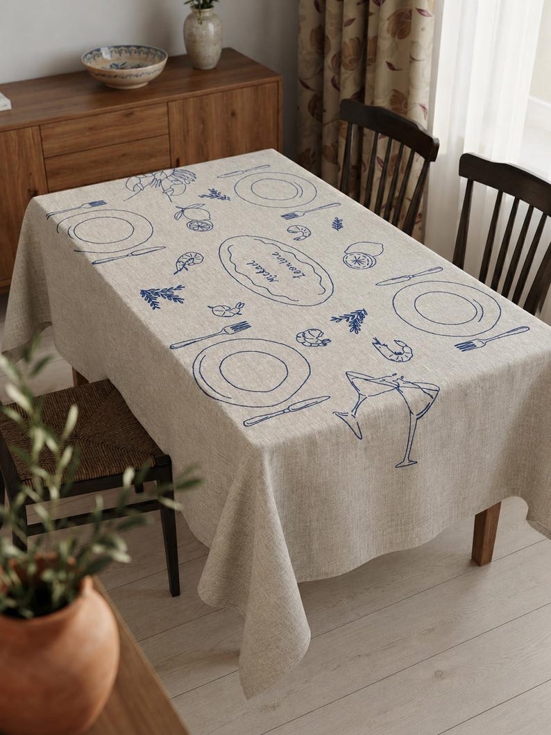 May include: A rectangular beige linen tablecloth featuring a blue line-art print of place settings, shrimp, lemons, herbs, and two clinking cocktail glasses. A central oval shape contains the handwritten names "Mickael" and "Leontine" in blue ink.