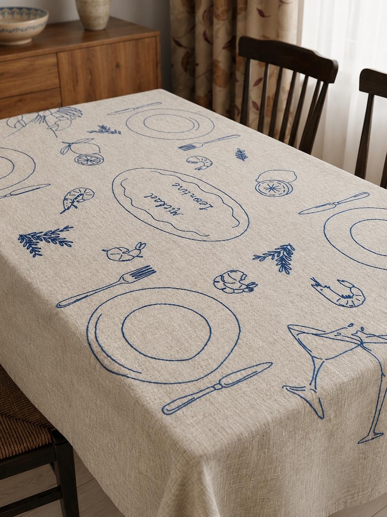 May include: A beige linen tablecloth featuring blue embroidered line art of plates, cutlery, shrimp, lemons, and two clinking glasses. The center oval embroidery displays the names Michael and Leontine in a cursive script font.