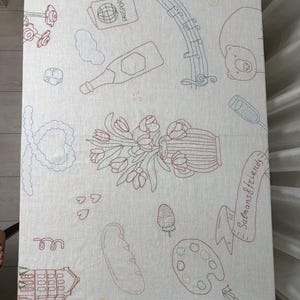 May include: A rectangular off-white fabric tablecloth featuring colorful embroidered line art illustrations including a passport, musical notes, a bottle, a vase of tulips, a baguette, a paint palette, and a building with the text Salmans & friends.