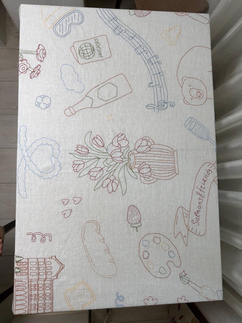 May include: A rectangular off-white fabric tablecloth featuring colorful embroidered line art illustrations including a passport, musical notes, a bottle, a vase of tulips, a baguette, a paint palette, and a building with the text Salmans & friends.