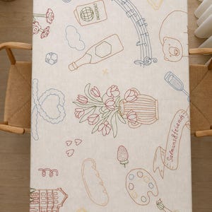 May include: A rectangular off-white tablecloth featuring colorful embroidered line art illustrations including a passport, musical notes, a bear, a vase of tulips, a paint palette, a building, and a banner with the text Salmans & friends.
