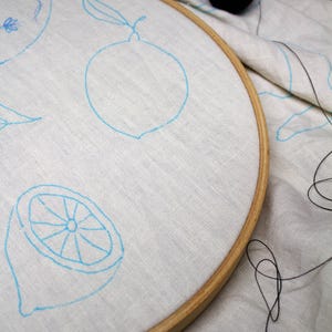 May include: A round wooden embroidery hoop holding a piece of off-white linen fabric. The fabric features light blue ink outlines of lemons and lemon slices. A small black spool of thread sits on the fabric outside the hoop with loose thread.