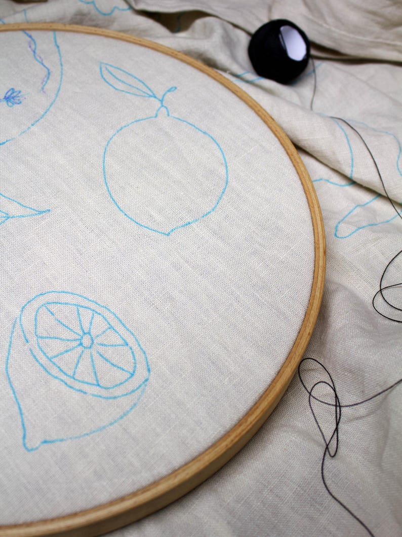 May include: A round wooden embroidery hoop holding a piece of off-white linen fabric. The fabric features light blue ink outlines of lemons and lemon slices. A small black spool of thread sits on the fabric outside the hoop with loose thread.