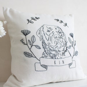 May include: Cream-colored square pillow with a black embroidered dog portrait. The dog is surrounded by floral and leaf designs. A banner below the dog reads "KIN".