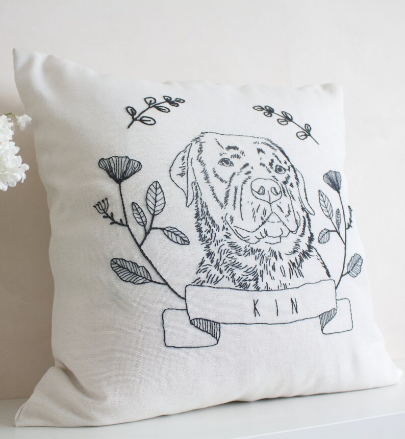 May include: Cream-colored square pillow with a black embroidered dog portrait. The dog is surrounded by floral and leaf designs. A banner below the dog reads "KIN".