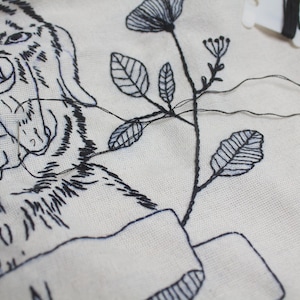 May include: Close-up of a white fabric with black embroidery. The embroidery depicts a tiger's face, flowers, and leaves. Black thread and a needle are visible, indicating an in-progress embroidery project. The design is detailed and intricate.