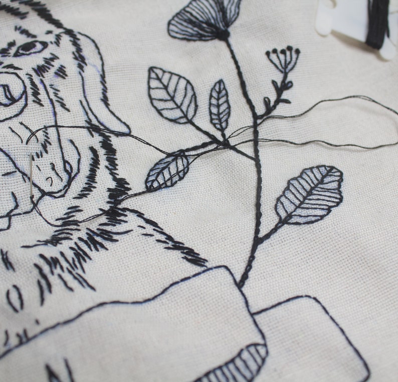 May include: Close-up of a white fabric with black embroidery. The embroidery depicts a tiger's face, flowers, and leaves. Black thread and a needle are visible, indicating an in-progress embroidery project. The design is detailed and intricate.