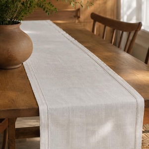 May include: A rectangular light beige table runner made of a woven fabric with a visible texture. The runner features a decorative hemstitch border detail along all four sides, creating a clean frame effect on the surface of the fabric.