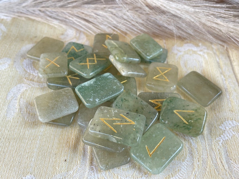 Puede incluir: Un conjunto de runas de piedra verde con s&iacute;mbolos dorados. Las runas tienen forma de rect&aacute;ngulos y est&aacute;n esparcidas sobre una superficie blanca.