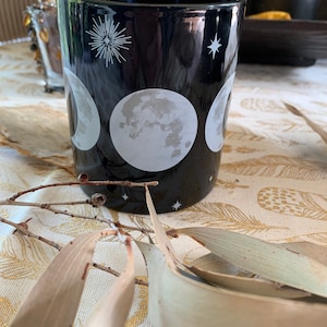 May include: Black ceramic mug with a white moon phases design. The mug features three different moon phases, a starburst, and a small star.