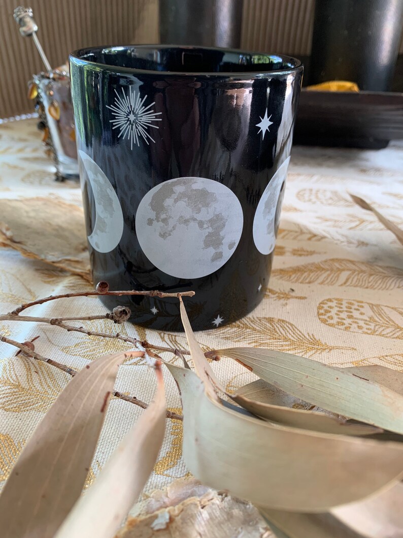 May include: Black ceramic mug with a white moon phases design. The mug features three different moon phases, a starburst, and a small star.