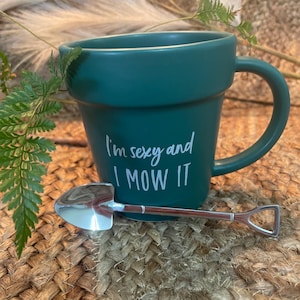 May include: A green ceramic mug shaped like a flower pot with the text "I'm sexy and I mow it" in white. A small silver shovel is resting on the rim of the mug.