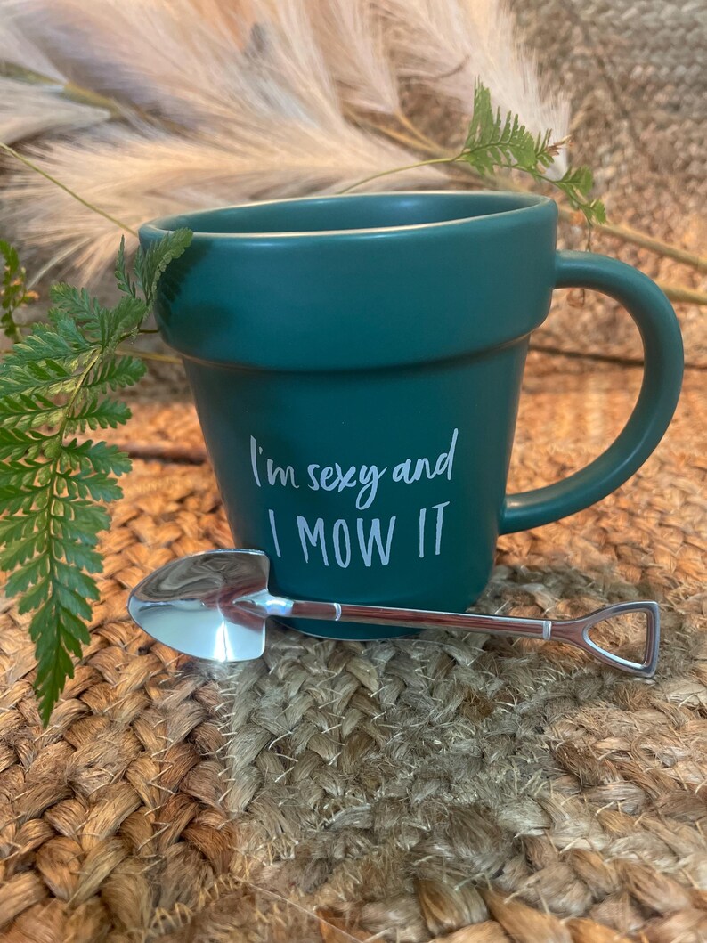 May include: A green ceramic mug shaped like a flower pot with the text "I'm sexy and I mow it" in white. A small silver shovel is resting on the rim of the mug.