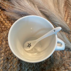 May include: A white ceramic teacup with a small white spoon inside. The spoon has a black five-pointed star design on it.