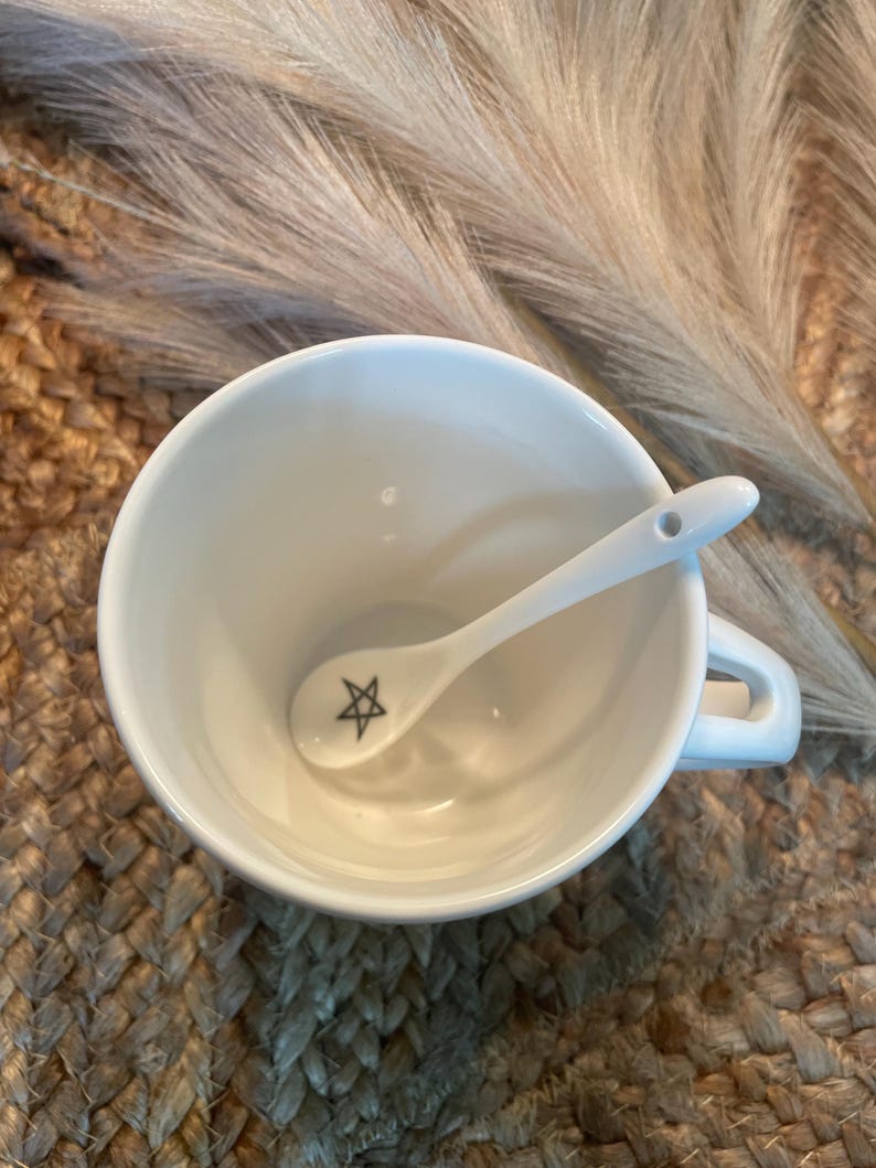 May include: A white ceramic teacup with a small white spoon inside. The spoon has a black five-pointed star design on it.