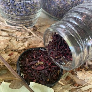 Op de afbeelding: Een close-up van een glazen pot gevuld met gedroogde hibiscusbloemen. De pot wordt in een kleine zwarte kom gegoten, waardoor de gedroogde rode en paarse bloemen eruit vallen.
