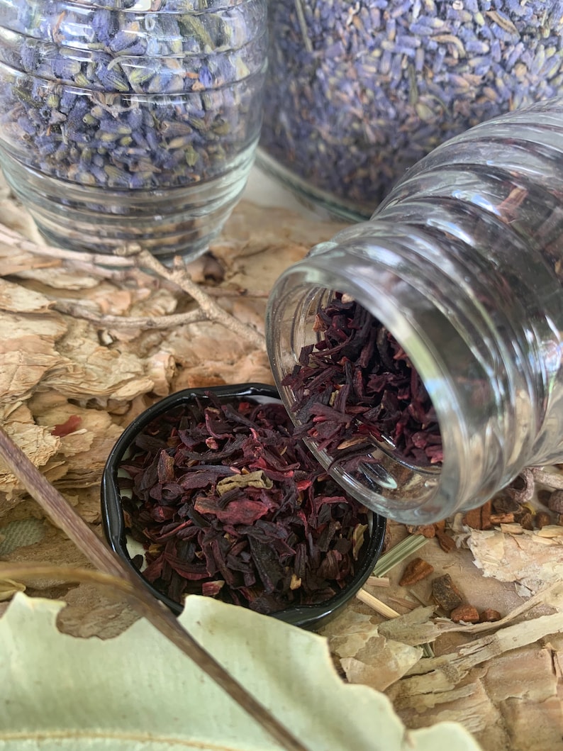Op de afbeelding: Een close-up van een glazen pot gevuld met gedroogde hibiscusbloemen. De pot wordt in een kleine zwarte kom gegoten, waardoor de gedroogde rode en paarse bloemen eruit vallen.