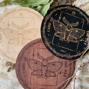K&ouml;nnte beinhalten: Drei runde Holz-Untersetzer mit einem Motten-Design und dem Text "Karte des Tages" auf jedem. Die Untersetzer sind in verschiedenen Holzt&ouml;nen: hellbraun, dunkelbraun und schwarz.