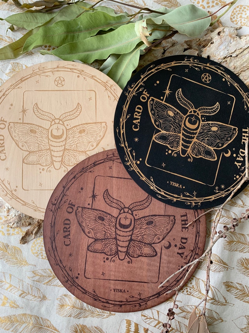 K&ouml;nnte beinhalten: Drei runde Holz-Untersetzer mit einem Motten-Design und dem Text "Karte des Tages" auf jedem. Die Untersetzer sind in verschiedenen Holzt&ouml;nen: hellbraun, dunkelbraun und schwarz.