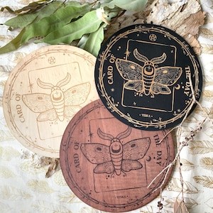 K&ouml;nnte beinhalten: Drei runde Holzschilder mit einem goldenen Mottivdesign und dem Text "Karte des Tages". Die Schilder sind in Schwarz, Naturholz und braunem Holz.