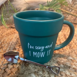 May include: A green ceramic mug with a white handle and the text "I'm sexy and I mow it". A small silver shovel is next to the mug.