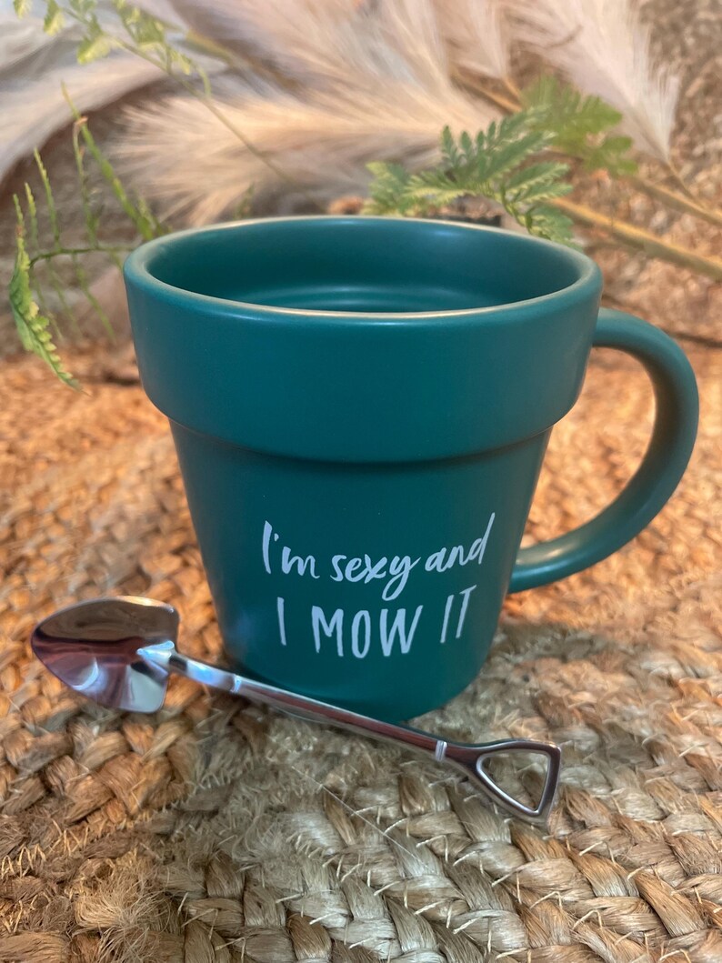 May include: A green ceramic mug with a white handle and the text "I'm sexy and I mow it". A small silver shovel is next to the mug.