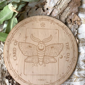 K&ouml;nnte beinhalten: Ein runder Holz-Kartenhalter mit einem floralen Rand und dem eingravierten Text "Karte des Tages". In der Mitte befindet sich eine detaillierte Illustration eines Nachtfalters mit einem Halbmond auf seinen Fl&uuml;geln.