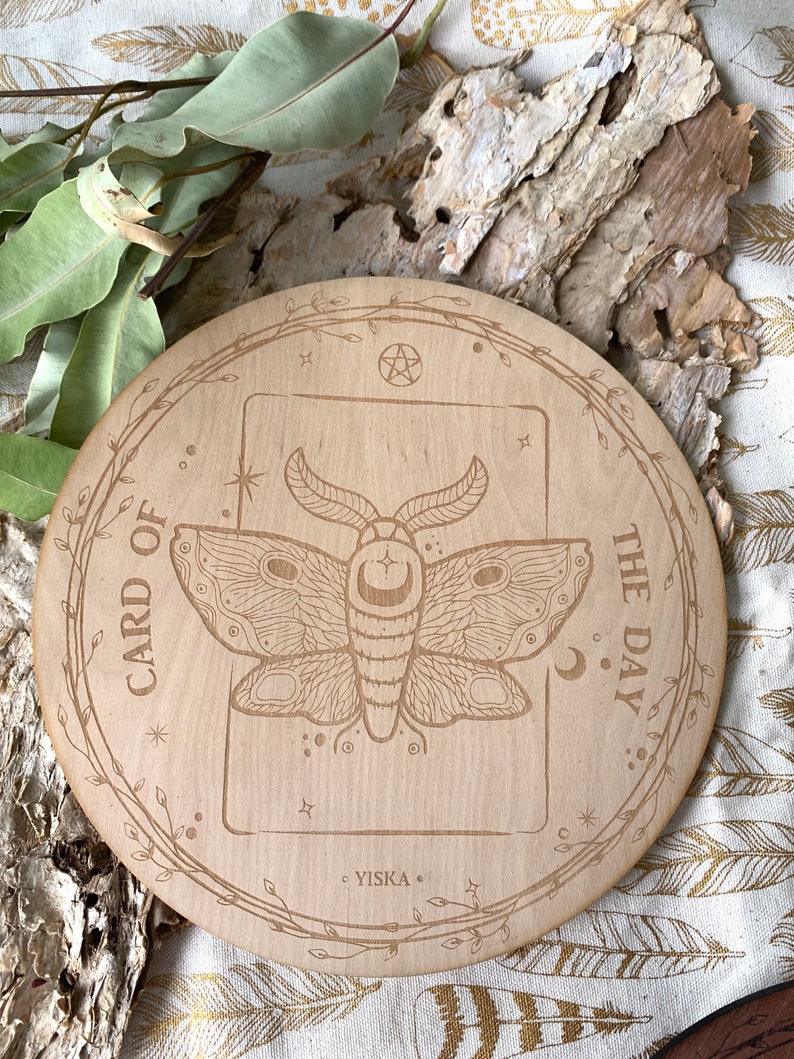 K&ouml;nnte beinhalten: Ein runder Holz-Kartenhalter mit einem floralen Rand und dem eingravierten Text "Karte des Tages". In der Mitte befindet sich eine detaillierte Illustration eines Nachtfalters mit einem Halbmond auf seinen Fl&uuml;geln.