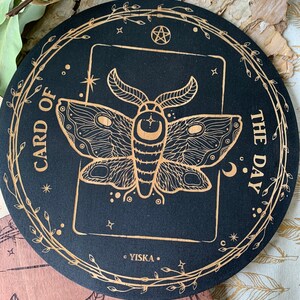 K&ouml;nnte beinhalten: Ein rundes Schild aus schwarzem Holz mit goldener Umrandung und einem goldenen Mottenmotiv im Inneren. Das Schild tr&auml;gt die Aufschrift "Karte des Tages" und hat einen Pentagramm und Sterne im Rand.