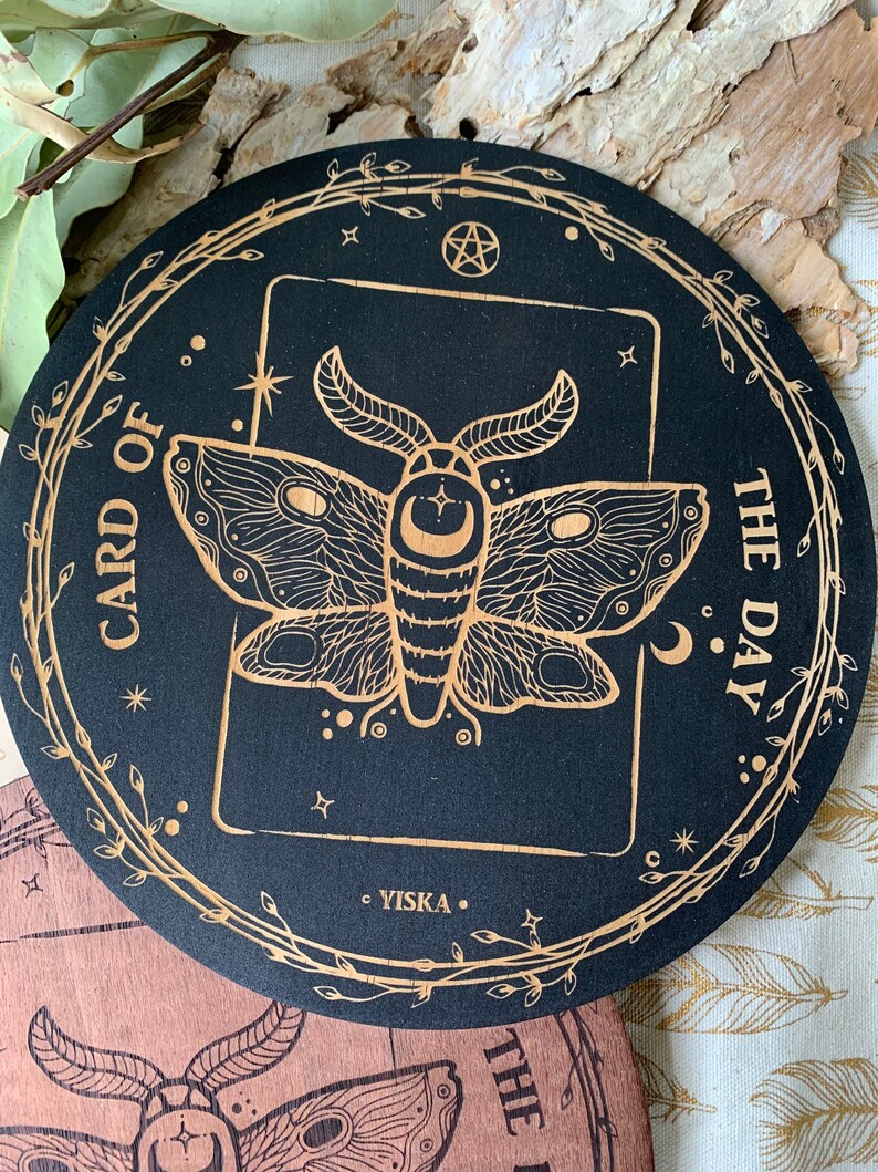 K&ouml;nnte beinhalten: Ein rundes Schild aus schwarzem Holz mit goldener Umrandung und einem goldenen Mottenmotiv im Inneren. Das Schild tr&auml;gt die Aufschrift "Karte des Tages" und hat einen Pentagramm und Sterne im Rand.