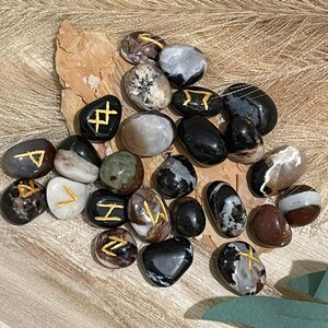 Puede incluir: Una colecci&oacute;n de piedras de &aacute;gata negra, blanca y marr&oacute;n con runas doradas pintadas sobre ellas.