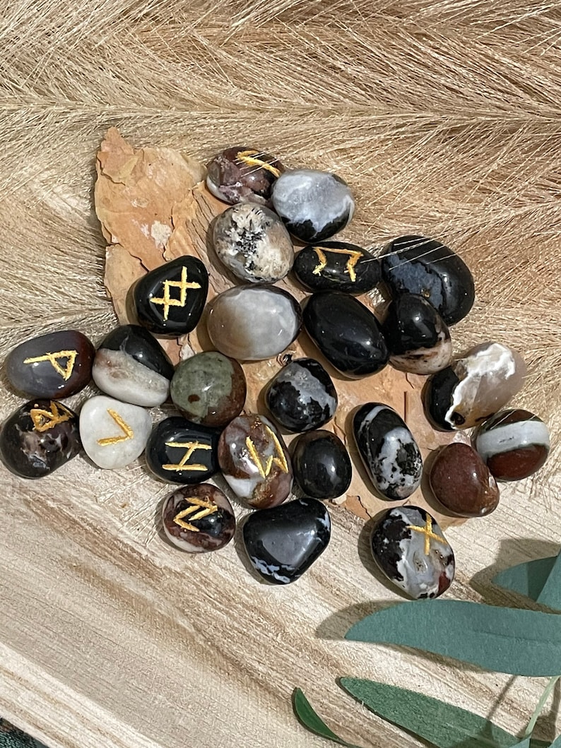 Puede incluir: Una colecci&oacute;n de piedras de &aacute;gata negra, blanca y marr&oacute;n con runas doradas pintadas sobre ellas.
