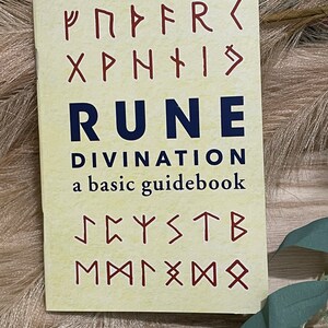 Puede incluir: Un libro amarillo con el t&iacute;tulo "Rune Divination: a basic guidebook" y runas en rojo y azul.