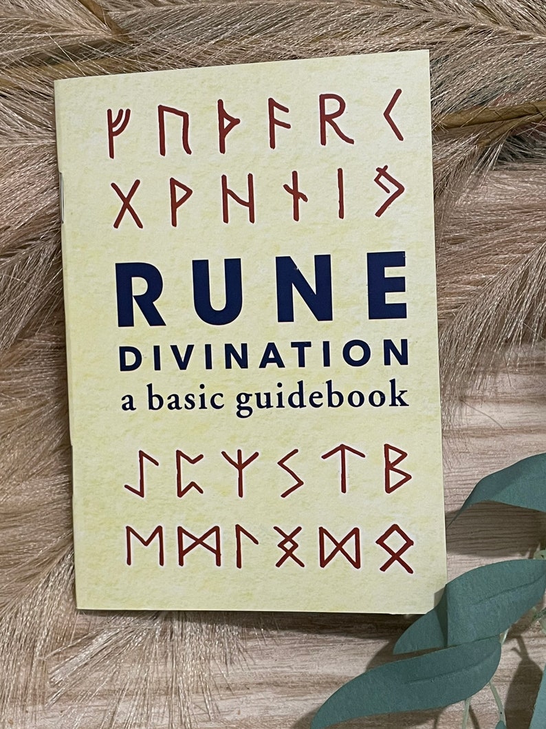 Puede incluir: Un libro amarillo con el t&iacute;tulo "Rune Divination: a basic guidebook" y runas en rojo y azul.