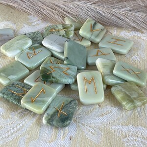Puede incluir: Un conjunto de runas de piedra verde y blanca con letras doradas. Las runas est&aacute;n en una pila sobre una superficie con un patr&oacute;n blanco y verde.