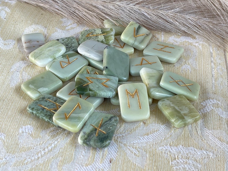 Puede incluir: Un conjunto de runas de piedra verde y blanca con letras doradas. Las runas est&aacute;n en una pila sobre una superficie con un patr&oacute;n blanco y verde.